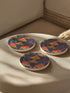 Round ceramic coaster featuring orange and yellow mushrooms with striped caps against a dark background with flowing purple lines in a psychedelic 70s style